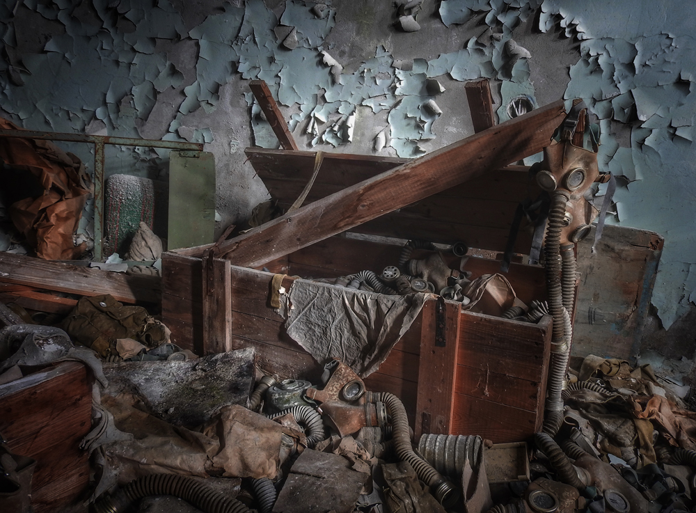 A broken wooden chest surrounded by scattered gas masks and debris occupies a dimly-lit room with peeling blue paint on the walls, suggesting abandonment and decay.