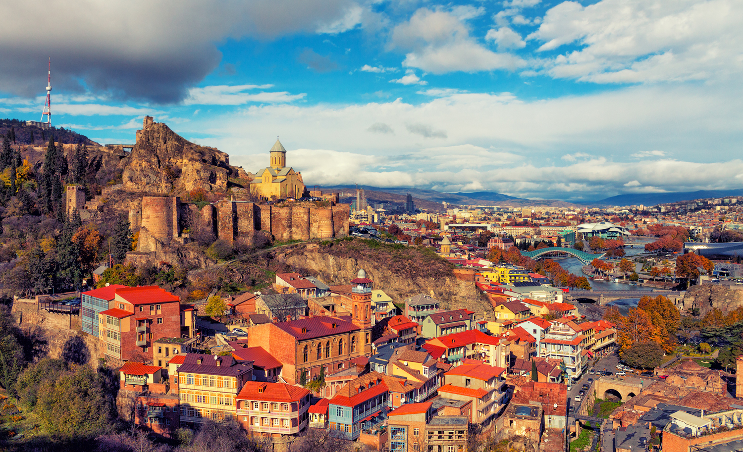 Fortress on a hill overlooks a city with colorful buildings, a winding river, and distant mountains under a partly cloudy sky, creating a vibrant urban landscape.