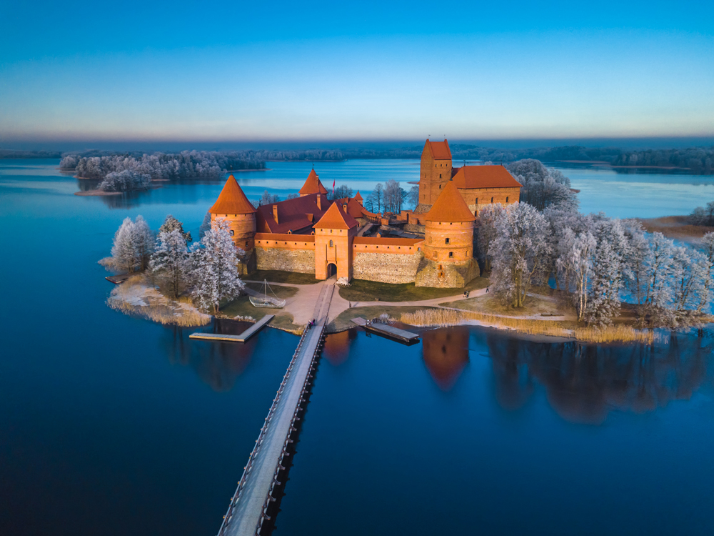 A historic brick castle with red rooftops stands on an island surrounded by a serene lake and frosty trees, connected to the shore by a long wooden bridge.