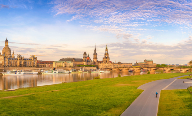 A cyclist rides along a riverside path with a backdrop of historical buildings and spired towers across a bridge, under a partly cloudy sky.