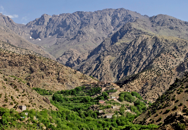 A small village rests amid lush green vegetation, situated within a broad valley. Towering rugged mountains form an imposing backdrop, with clear blue skies overhead.
