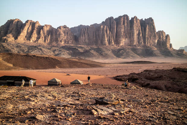 Rocky desert landscape with towering cliffs in the background. A few tents sit on the desert floor. Two people stand overlooking the scenery, with mountains towering in the distance.