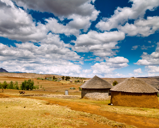 Two round thatched huts stand in an open, grassy field under a partly cloudy blue sky, with distant hills and scattered trees in the rural landscape.