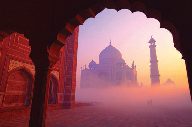 The Taj Mahal stands majestically in the misty sunrise, viewed through an ornate arched doorway. Silhouettes of visitors and a tall minaret enhance the atmospheric scene.