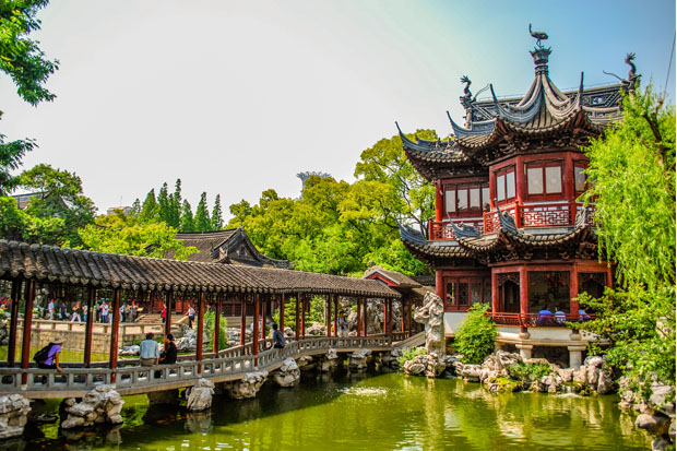 Ornate traditional Chinese building stands beside a scenic pond. Visitors walk along a covered wooden bridge, surrounded by lush greenery and trees, creating a tranquil garden setting.
