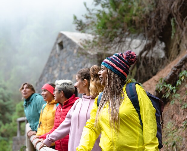 Group of five people stand smiling and leaning on a wooden fence, dressed in colorful jackets, amidst foggy forest surroundings with a stone building in the background.