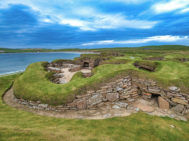 Stone ruins form ancient structures nestled on an elevated grassy terrain beside a calm sea, under a sky filled with scattered clouds.