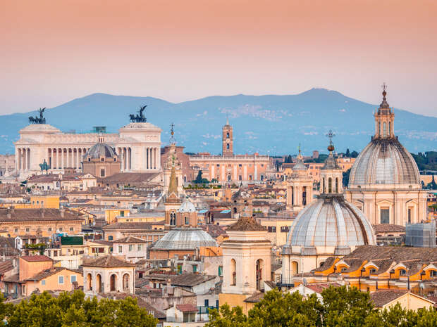 Cityscape featuring historic buildings with domes and statues under a pastel sky. Mountains in the background provide a natural backdrop. Rooftops and trees fill the foreground.