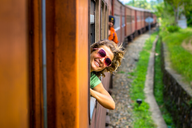 A person with sunglasses leans out of a train window, smiling joyfully; the train moves through a lush, green countryside, with another passenger visible in the background.