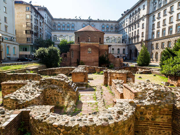 Brick rotunda stands centrally, surrounded by ancient stone ruins, in a courtyard bordered by grand, multi-story buildings. Greenery adds contrast, indicating a mix of history and urban architecture.