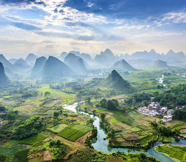 Lush green fields with a winding river cross through a valley surrounded by misty, jagged mountains under a partly cloudy sky. A cluster of small buildings sits to the right.