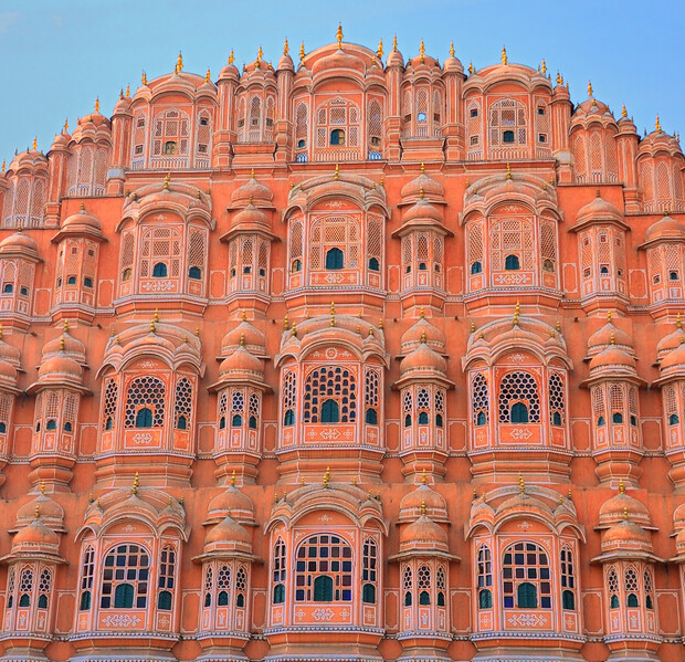 A pink sandstone facade with numerous ornate windows, showcasing intricate lattice designs and domed arches, stands against a blue sky, creating a striking architectural display.