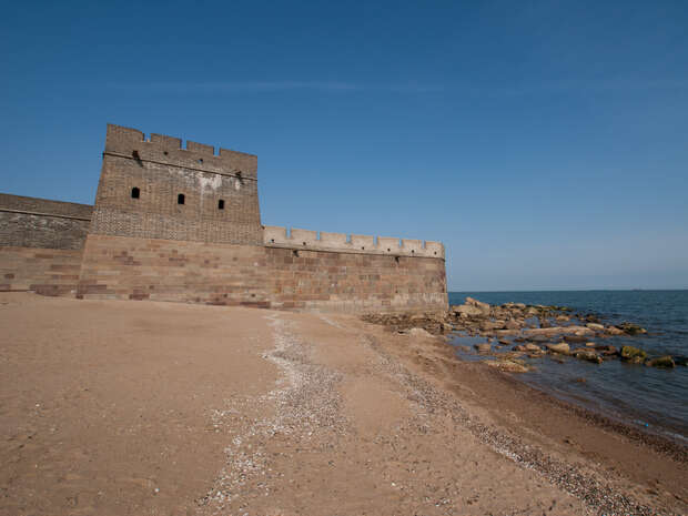 A stone fortification extends along a sandy beach, meeting the sea. The structure features battlements and rectangular towers, while rocks dot the shoreline under a clear blue sky.