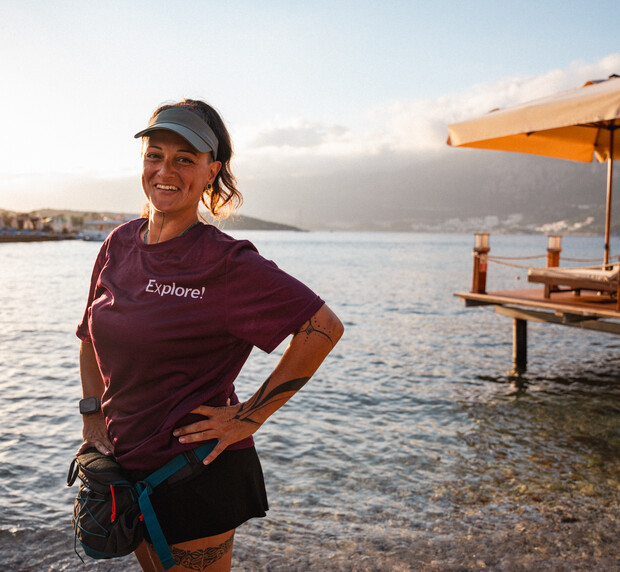 A person stands smiling on a rocky shoreline, wearing a purple shirt with "Explore!" printed on it, with a visor and waist bag. A dock and umbrella visible nearby under a cloudy sky.