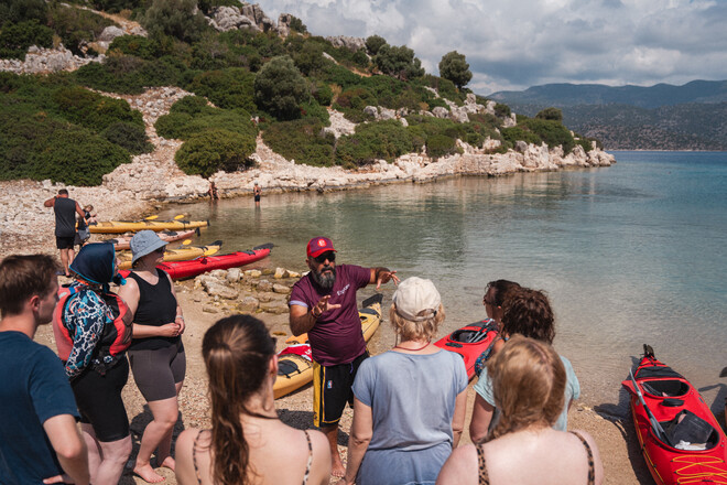 A group of people listens to a guide gesturing with hands on a pebbled beach, near colorful kayaks, against a backdrop of hilly greenery and calm sea.