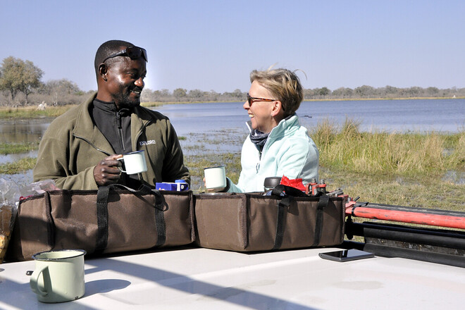 Two people holding mugs, smiling and chatting, stand by a lakeside. They are surrounded by bags on a tabletop positioned on a vehicle. The lake and tall grass are in the background.