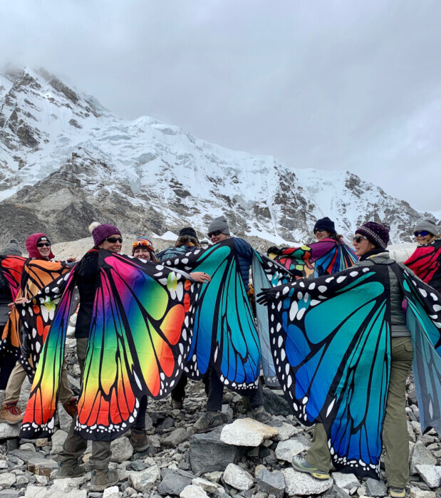 People wearing colorful butterfly wing costumes stand on rocky terrain. In the background, a snow-covered mountain looms under an overcast sky. They face each other, smiling and chatting.