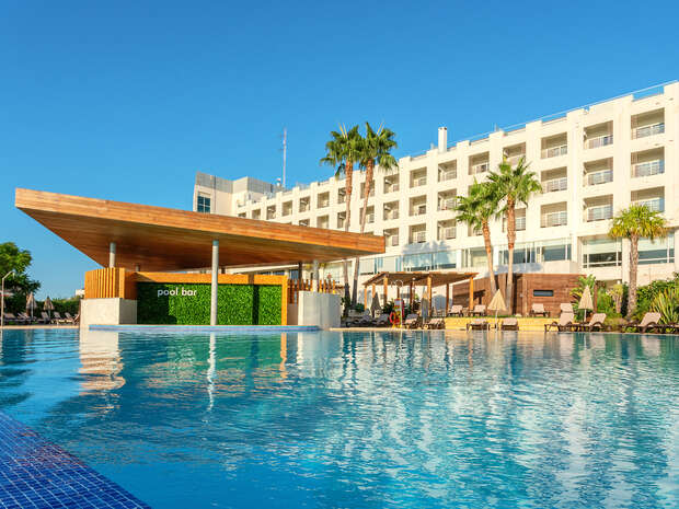 A wooden-roofed pool bar labeled "pool bar" stands adjacent to a large pool, surrounded by lounge chairs and palm trees, in front of a white multi-story hotel under a clear blue sky.
