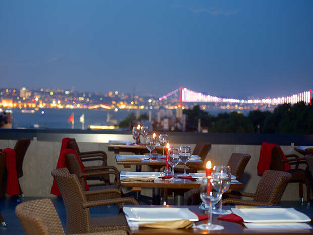 Wicker dining chairs surround candlelit tables on an outdoor terrace. In the background, a city skyline glows with lights, featuring a prominently lit bridge against a twilight sky.