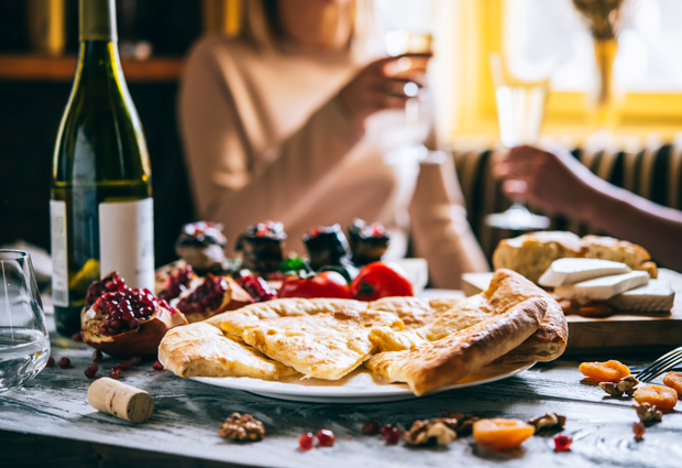 A plate of sliced flatbread sits on a table with pomegranates, nuts, and cheese nearby, while two people in the background hold wine glasses in a warmly lit room.