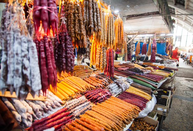 Colorful churchkhela candies hang and lie on tables in a bustling indoor market, with various vibrant hues of red, orange, and brown, surrounded by vendors and shoppers.
