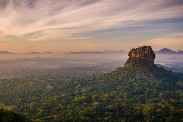 A prominent rock formation rises above lush green forests, under a vast sky with scattered clouds, creating a serene and majestic landscape.