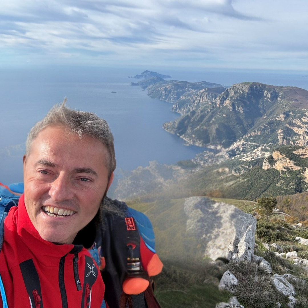 A person in a red and blue jacket smiles while standing on a cliffside. The background displays a scenic coastal landscape with cliffs and the sea extending to the horizon.