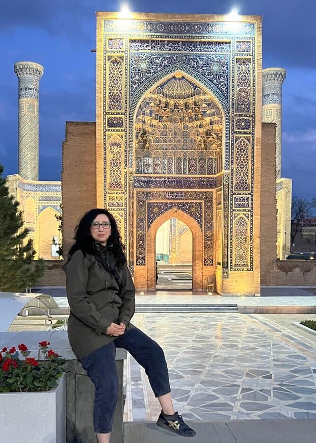 A person sits on a ledge, hands clasped, against an ornately decorated, illuminated arch at night. The background includes tiled surfaces, trees, and tall, cylindrical minarets.