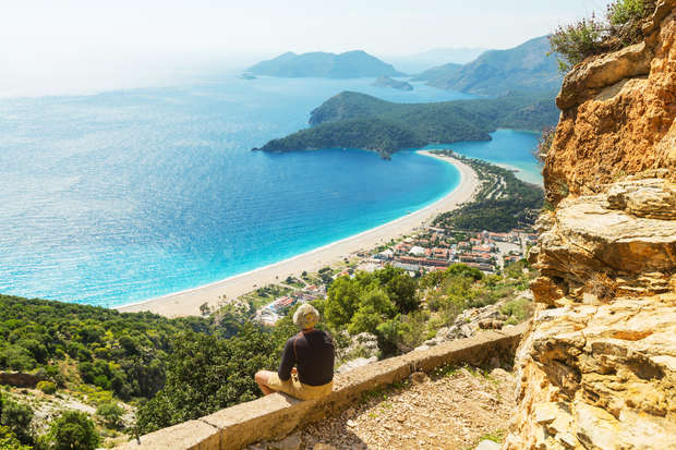 Person sits on a stone ledge, gazing at a turquoise sea bay and distant hills, surrounded by lush greenery and rocky formations, with a small town below.
