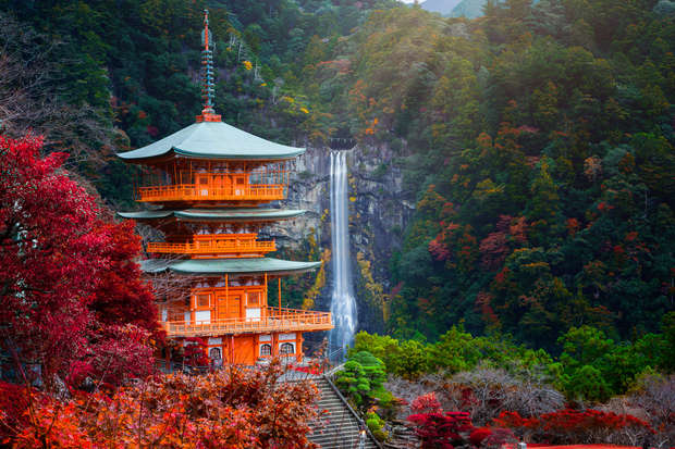A multi-tiered pagoda stands amidst vibrant autumn foliage with a cascading waterfall in the background, surrounded by lush forested hills.