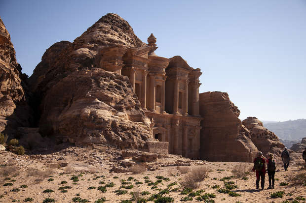 A large ancient stone structure is carved into rocky cliffs under a clear blue sky. Three people walk across the sandy, shrub-dotted ground, highlighting its historical and desert setting.