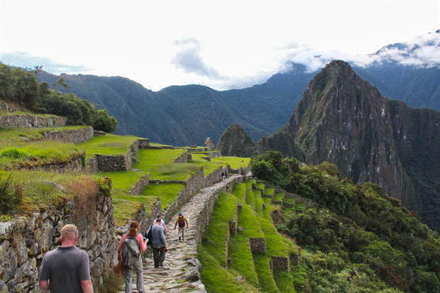 People walk along an ancient stone path amidst terraced green landscapes, mountains, and cloudy skies, reminiscent of a historic site, creating a sense of exploration and wonder.