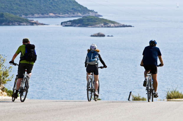 Three cyclists ride down a road toward a blue sea, with hilly islands in the background under a clear sky, creating a scenic coastal journey.