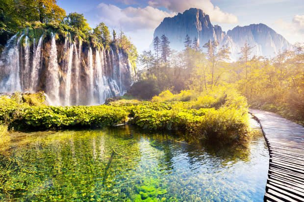Waterfalls cascade over a rocky cliff into a clear pond. A wooden path curves alongside the water, surrounded by vibrant greenery and distant, hazy mountains under a bright sky.