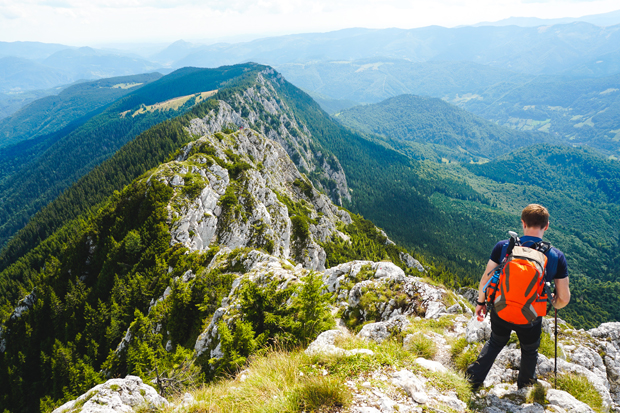 A hiker with an orange backpack carefully walks along a mountain ridge, surrounded by lush green forests and distant rolling hills under a partly cloudy sky.