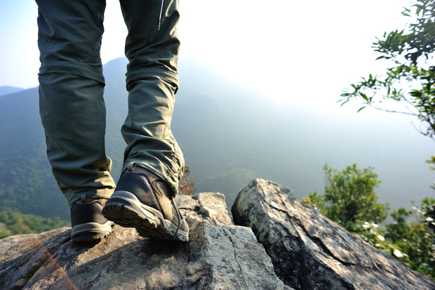 Hiking boots stand firmly on rocky terrain. The person appears to be looking out over a misty, mountainous landscape, surrounded by greenery and distant peaks, under a bright sky.