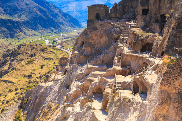 Rock-cut caves with pathways and railings ascend a steep cliffside. In the background, a lush valley and rugged mountains stretch into the distance, under a clear blue sky.
