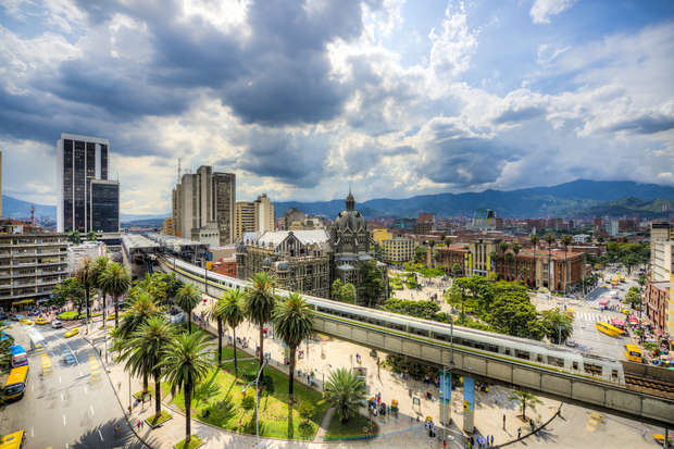 A bustling cityscape features prominent historical architecture surrounded by tall modern buildings. A train moves along an elevated track through palm trees and a vibrant urban environment under a partly cloudy sky.