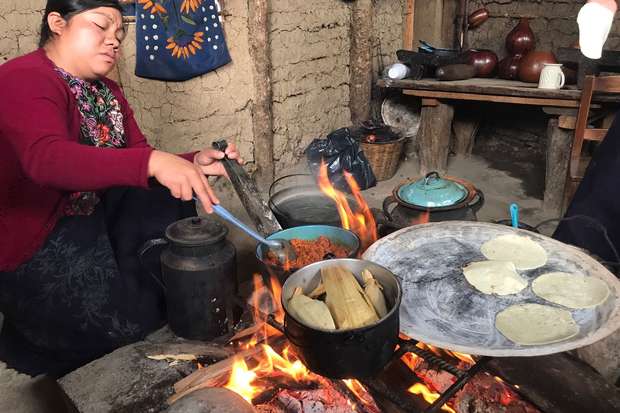 A woman cooks tortillas on a hot griddle over an open flame in a rustic kitchen. She stirs a nearby pot while surrounded by cookware and traditional items in an earthen-walled room.