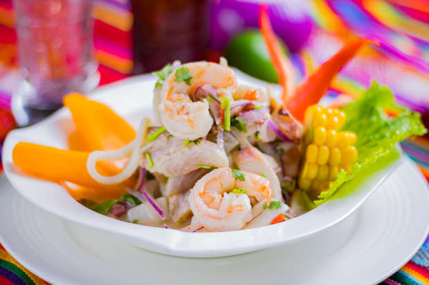 A bowl of ceviche with shrimp and fish is garnished with cilantro, red onion, corn, and bell pepper slices, set on a white plate. The colorful background suggests a festive dining table.