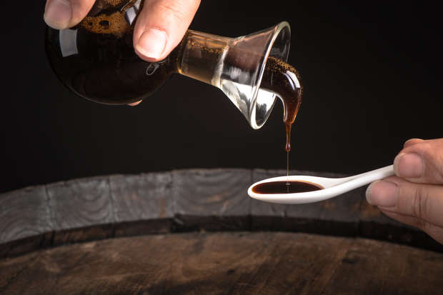 A clear glass bottle pours dark liquid into a white ceramic spoon, held over a wooden barrel, set against a dark background.