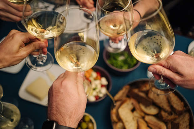 Glasses filled with white wine clink together above a table. Surrounding items include plates of sliced bread, cheese, salad, and olives, suggesting a celebratory meal setting.
