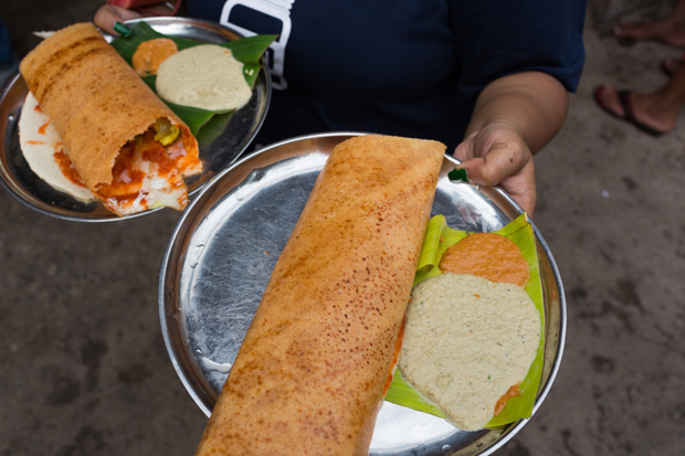 A person holds two plates of dosa, with sambar and chutney, on a street. The dosas are large and golden brown, served on metal plates lined with banana leaves.