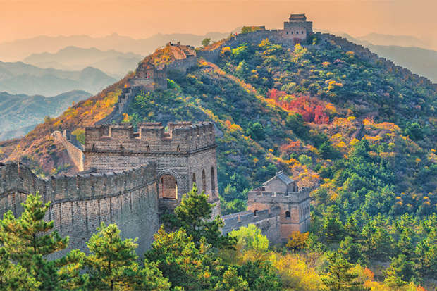 A section of the Great Wall winds over a lush, colorful hillside, surrounded by vibrant autumn foliage and distant hazy mountains under a soft, orange sky.
