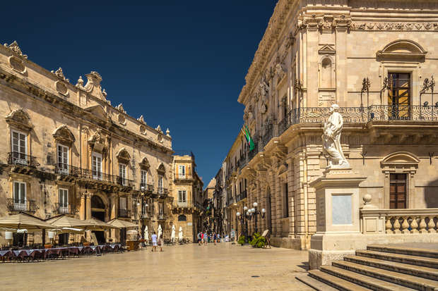 Historic stone buildings line a sunlit square, featuring ornate balconies and a statue on a pedestal. People walk or sit at an outdoor café, flags hanging from façades under a clear blue sky.