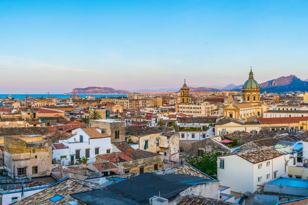 Rooftops stretch across an urban landscape, dominated by historic buildings and church domes. In the background, mountains meet a clear blue sky, with a glimpse of the sea on the horizon.