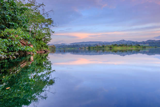 A calm lake reflects trees and a colorful sky, surrounded by lush greenery and distant hills at dusk, creating a tranquil natural scene.