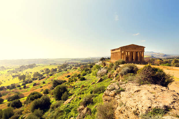 Ancient temple sits atop a lush hill, basking in sunlight, surrounded by expansive countryside with rolling fields and scattered trees, under a clear blue sky.