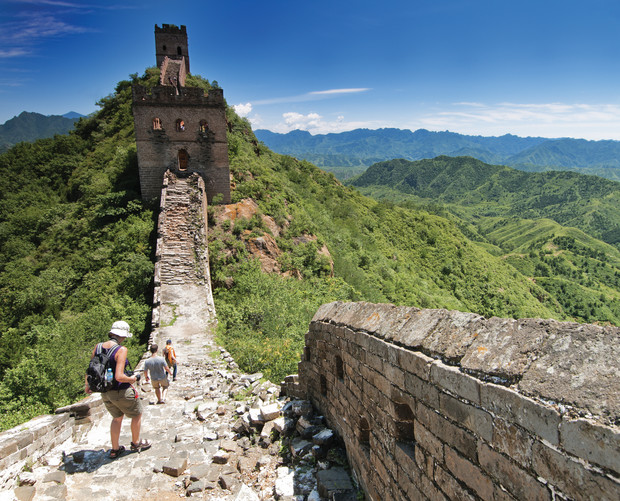 Tourists walk along a rugged section of the Great Wall of China, with a watchtower visible ahead. Lush green mountains surround the ancient stone structure under a clear blue sky.