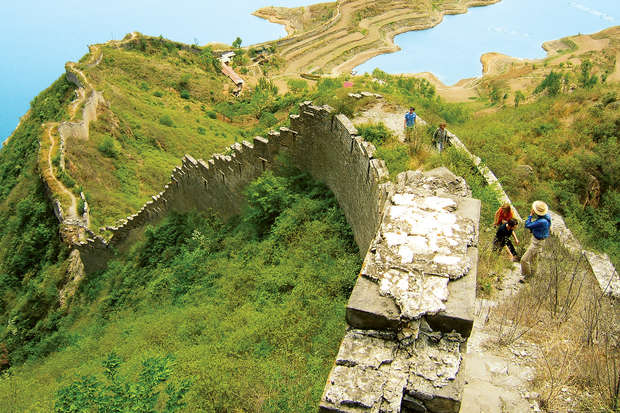 A section of the Great Wall extends through green hills, with people walking along its crumbling path. In the background, terraced landscapes meet a serene, blue body of water.